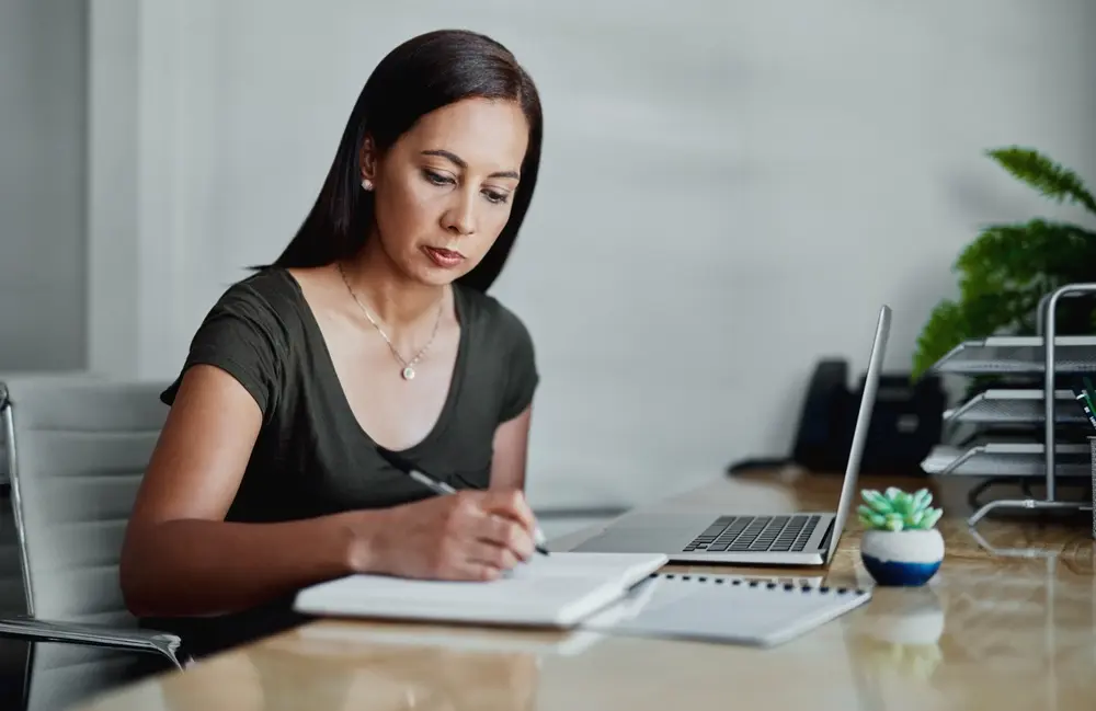 Office setup with a laptop and a businesswoman writing in a notebook.